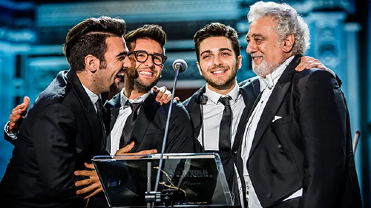 Czterech mężczyzn w garniturach, w tym legendarny tenor, który ma najwyższy głos męski, śmieje się na scenie.