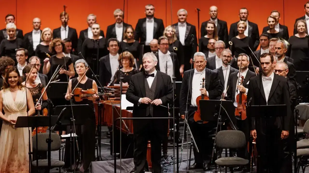 Orkiestra i chór przygotowują się do wykonania pieśni Moniuszki. Dyrygent stoi na podwyższeniu, muzycy trzymają instrumenty.