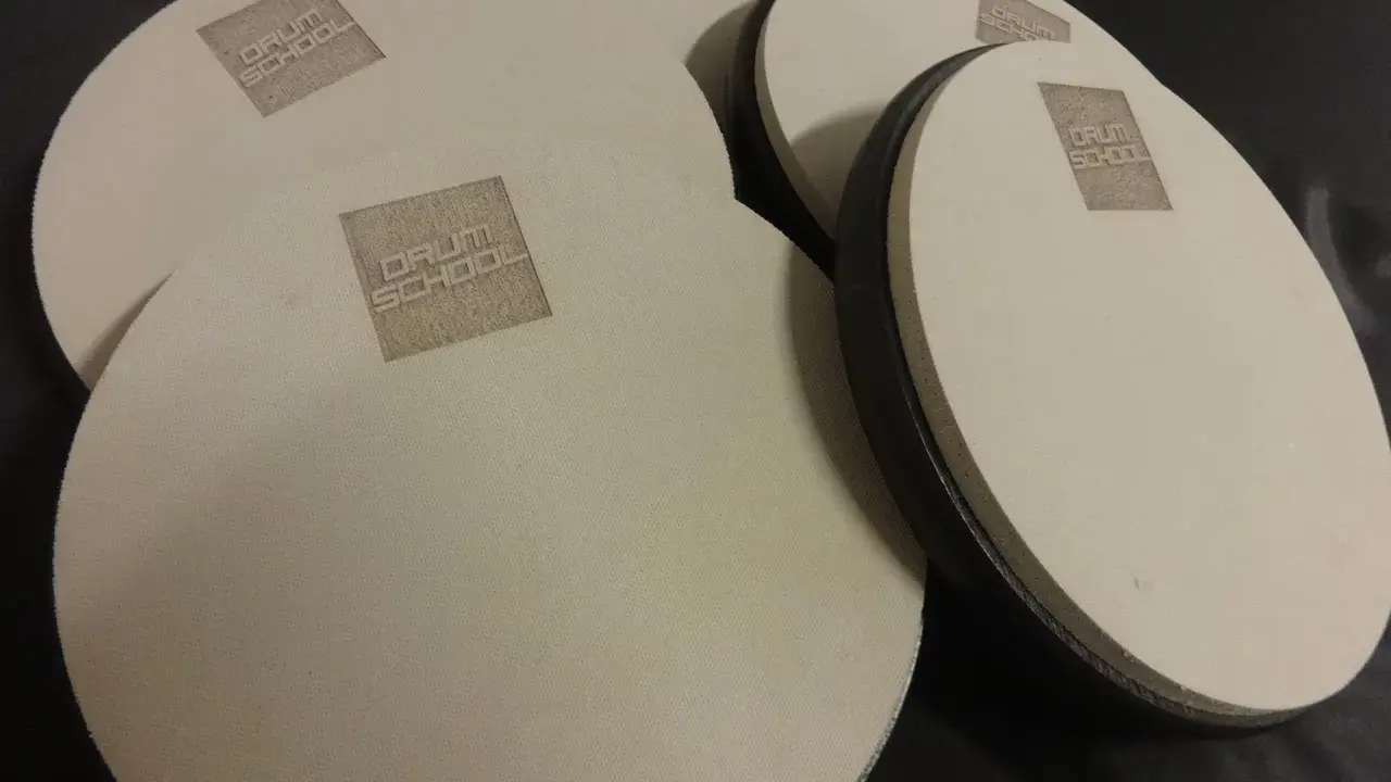 Kilka okrągłych, beżowych padów perkusyjnych z logo "DRUM SCHOOL" leży na czarnym tle.