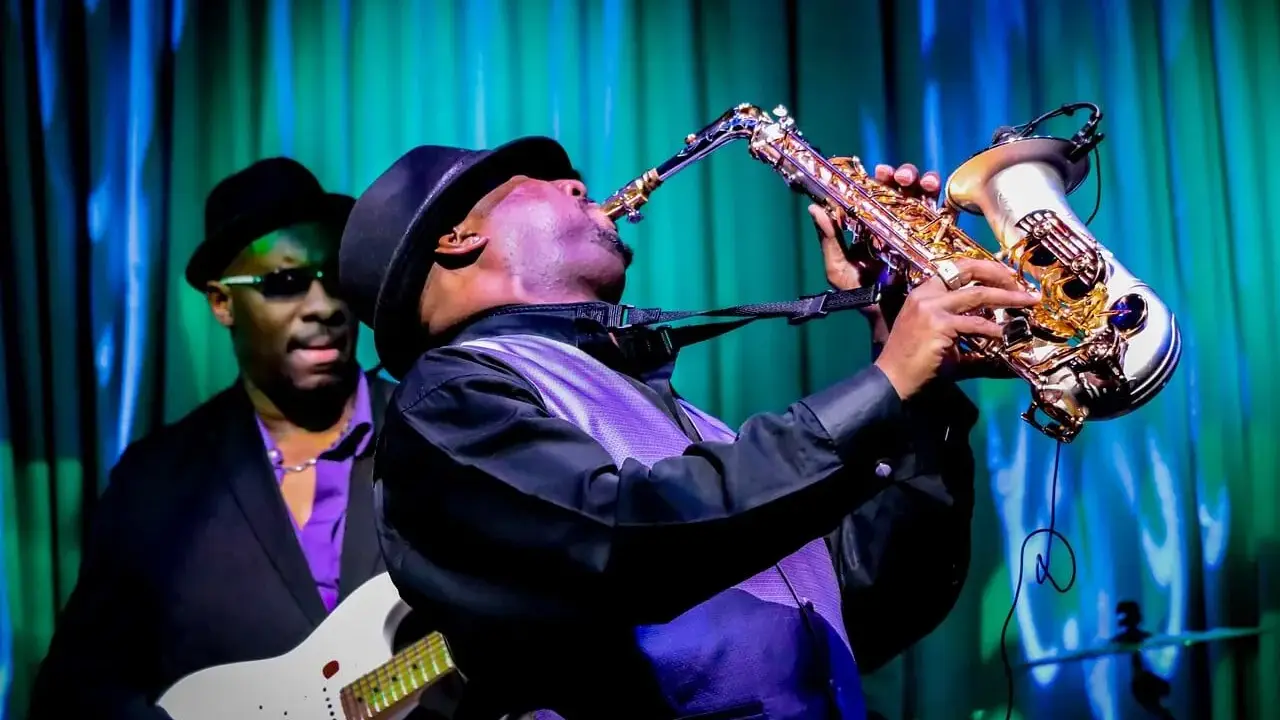 Muzyk gra na saksofonie, tworząc nowoczesny jazz. W tle widać gitarzystę.