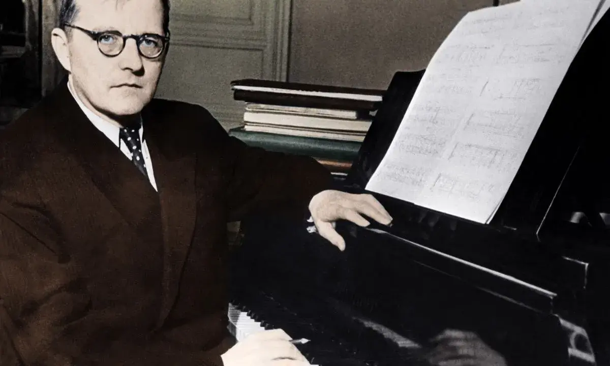 Dmitri Shostakovich, w okularach i garniturze, siedzi przy fortepianie, z nutami przed sobą.
