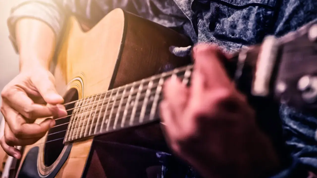 Ręce grające na gitarze akustycznej. Zbliżenie na palce naciskające struny i kostkę. Idealny moment, by zacząć naukę, jak nauczyć się grać na gitarze.