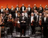 Orkiestra i chór przygotowują się do wykonania pieśni Moniuszki. Dyrygent stoi na podwyższeniu, muzycy trzymają instrumenty.