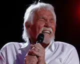 Kenny Rogers z zamkniętymi oczami śpiewa do mikrofonu, przypominając swoje niezapomniane utwory.