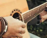 Dłoń z bransoletką z plecionej skóry gra na gitarze akustycznej.