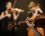 Dłonie artysty grają na skrzypcach, tworząc wariację na temat klasycznej melodii. W tle widać innych muzyków.