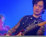 Jan Borysewicz gra na czerwonej gitarze elektrycznej, jego rękawy zdobią tatuaże.