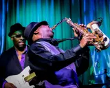 Muzyk gra na saksofonie, tworząc nowoczesny jazz. W tle widać gitarzystę.