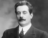 Portret Giacomo Pucciniego, kompozytora operowego, z charakterystycznymi wąsami i w eleganckim garniturze.