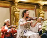 Koncert w stylu epoki Mozarta. W centrum uśmiechnięta kobieta grająca na skrzypcach, przypominająca żonę Mozarta.
