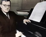 Dmitri Shostakovich, w okularach i garniturze, siedzi przy fortepianie, z nutami przed sobą.