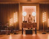 Dj perkusista w eleganckim stroju, z mikrofonem w dłoni, gotowy do zabawy. Obok zestaw perkusyjny i sceniczne oświetlenie.