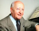 Uśmiechnięty Witold Lutosławski w garniturze przy fortepianie, z nutami na pulpicie.