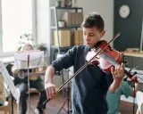 Chłopiec gra na skrzypcach, otoczony innymi dziećmi ćwiczącymi muzykę. Wszyscy skupieni, panuje **cicho w muzyce**, czekając na wspólne wykonanie.