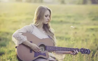 Dziewczyna gra na gitarze, tworząc piękny riff. Słońce delikatnie oświetla jej blond włosy i koronkową sukienkę.