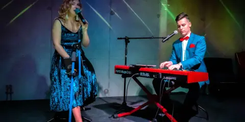 Wokalistka w niebieskiej sukni i klawiszowiec w garniturze tworzą duet. Ich występ to przykład kameralnych zespołów wokalnych.