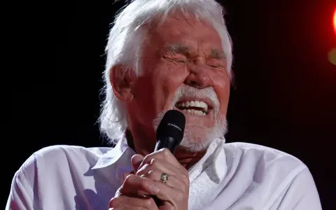 Kenny Rogers z zamkniętymi oczami śpiewa do mikrofonu, przypominając swoje niezapomniane utwory.