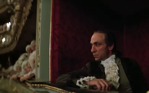 Scena z filmu o Mozarcie. Mężczyzna w stroju z epoki, w loży teatralnej, z widownią w tle.