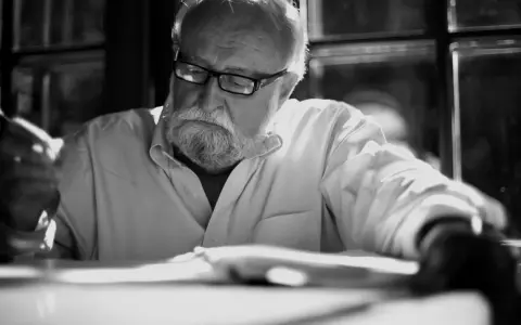 Krzysztof Penderecki w okularach, z siwą brodą, pochylony nad książką, z piórem w dłoni.