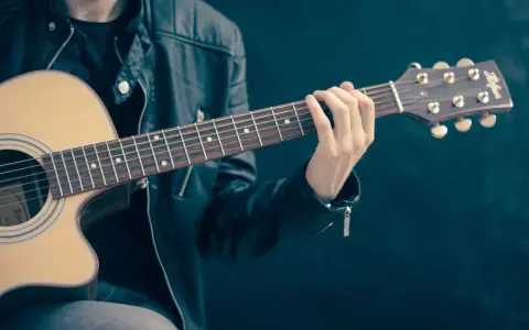 Młody mężczyzna w skórzanej kurtce gra na gitarze akustycznej. Dłoń na gryfie pokazuje, czym się gra na gitarze.