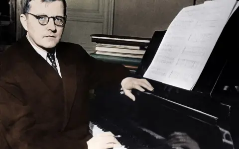 Dmitri Shostakovich, w okularach i garniturze, siedzi przy fortepianie, z nutami przed sobą.