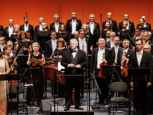 Orkiestra i chór przygotowują się do wykonania pieśni Moniuszki. Dyrygent stoi na podwyższeniu, muzycy trzymają instrumenty.