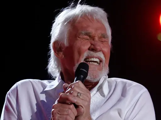 Kenny Rogers z zamkniętymi oczami śpiewa do mikrofonu, przypominając swoje niezapomniane utwory.