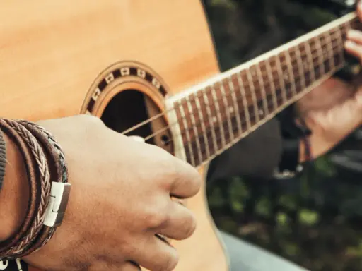 Dłoń z bransoletką z plecionej skóry gra na gitarze akustycznej.