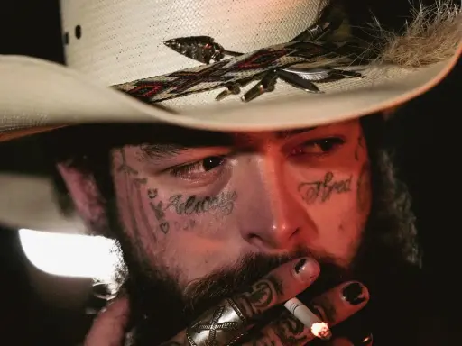 Amerykańskie gwiazdy muzyki country, jak Post Malone, w kowbojskim kapeluszu, z tatuażami na twarzy, palący papierosa.