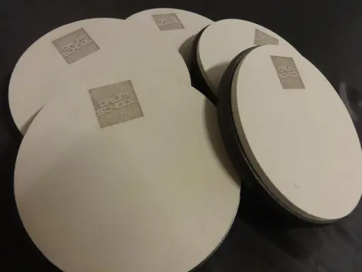 Kilka okrągłych, beżowych padów perkusyjnych z logo "DRUM SCHOOL" leży na czarnym tle.