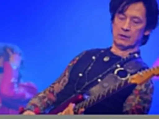 Jan Borysewicz gra na czerwonej gitarze elektrycznej, jego rękawy zdobią tatuaże.