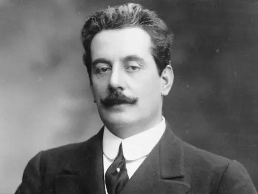 Portret Giacomo Pucciniego, kompozytora operowego, z charakterystycznymi wąsami i w eleganckim garniturze.