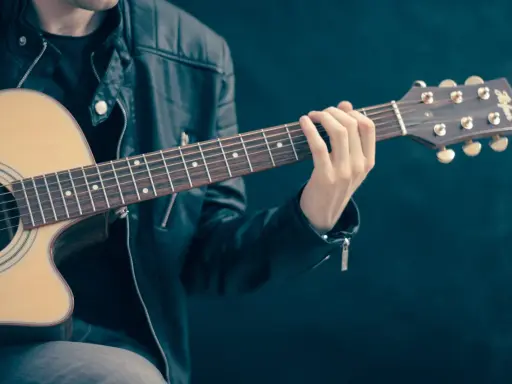 Młody mężczyzna w skórzanej kurtce gra na gitarze akustycznej. Dłoń na gryfie pokazuje, czym się gra na gitarze.
