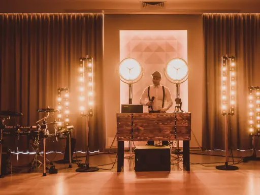 Dj perkusista w eleganckim stroju, z mikrofonem w dłoni, gotowy do zabawy. Obok zestaw perkusyjny i sceniczne oświetlenie.