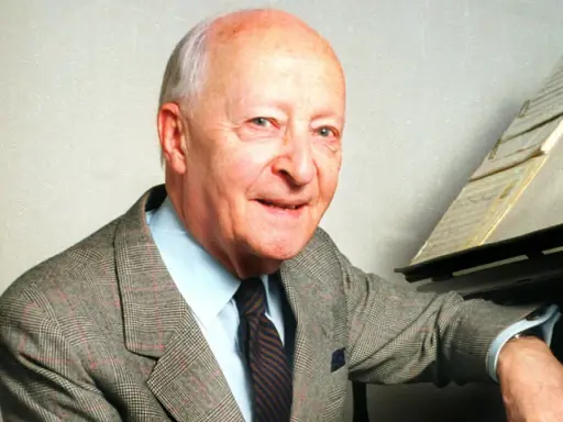Uśmiechnięty Witold Lutosławski w garniturze przy fortepianie, z nutami na pulpicie.