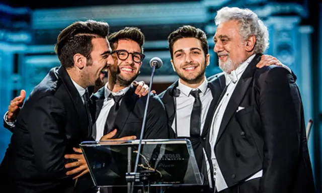 Czterech mężczyzn w garniturach, w tym legendarny tenor, który ma najwyższy głos męski, śmieje się na scenie.