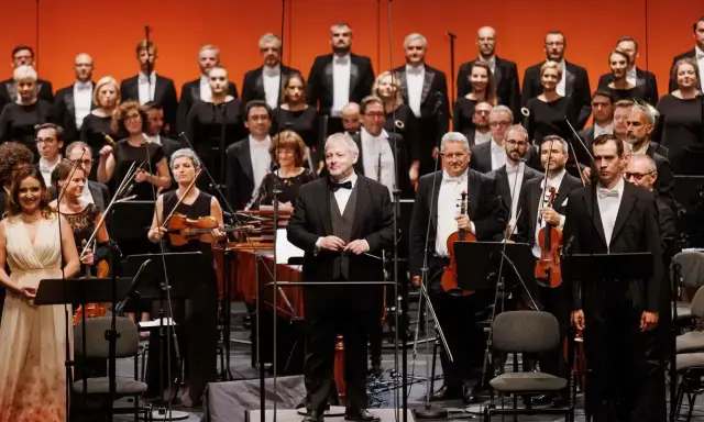 Orkiestra i chór przygotowują się do wykonania pieśni Moniuszki. Dyrygent stoi na podwyższeniu, muzycy trzymają instrumenty.