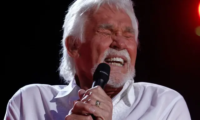 Kenny Rogers z zamkniętymi oczami śpiewa do mikrofonu, przypominając swoje niezapomniane utwory.