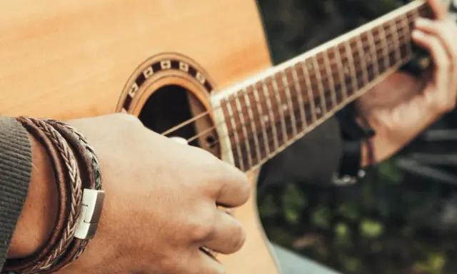 Dłoń z bransoletką z plecionej skóry gra na gitarze akustycznej.
