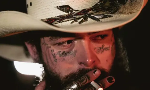Amerykańskie gwiazdy muzyki country, jak Post Malone, w kowbojskim kapeluszu, z tatuażami na twarzy, palący papierosa.