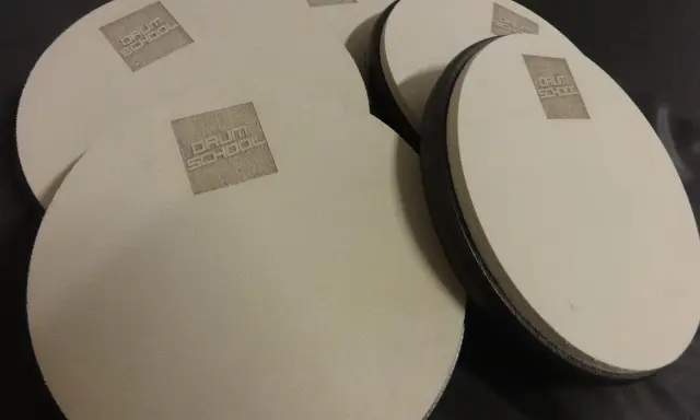 Kilka okrągłych, beżowych padów perkusyjnych z logo "DRUM SCHOOL" leży na czarnym tle.