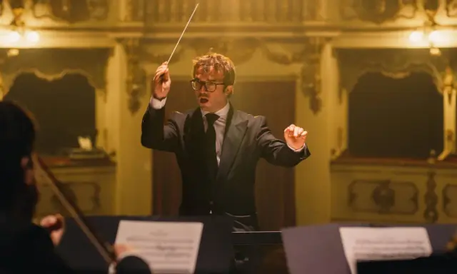 Dyrygent w garniturze prowadzi orkiestrę, tworząc muzykę godną najlepszych polskich kompozytorów muzyki filmowej.