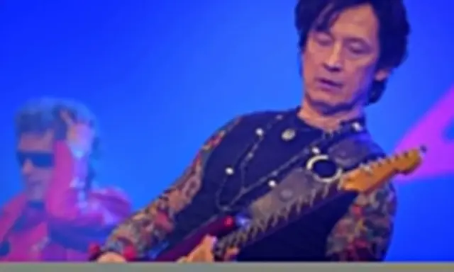 Jan Borysewicz gra na czerwonej gitarze elektrycznej, jego rękawy zdobią tatuaże.