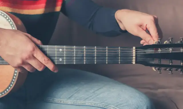 Mężczyzna stroi gitarę 12 strunową, kręcąc kluczem przy główce instrumentu.