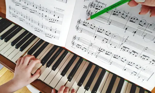Dziecko uczy się grać na pianinie, ćwicząc nuty z podręcznika. Zielony ołówek wskazuje fragment partytury, co ułatwia naukę gry na instrumentach dla dzieci.