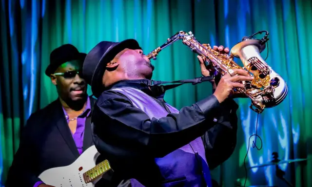Muzyk gra na saksofonie, tworząc nowoczesny jazz. W tle widać gitarzystę.