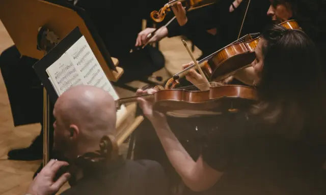 Skupieni muzycy grają na skrzypcach podczas części koncertu bez solisty. Nutki na pulpicie wskazują melodię.