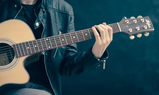 Młody mężczyzna w skórzanej kurtce gra na gitarze akustycznej. Dłoń na gryfie pokazuje, czym się gra na gitarze.