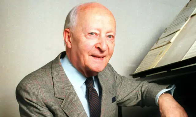Uśmiechnięty Witold Lutosławski w garniturze przy fortepianie, z nutami na pulpicie.