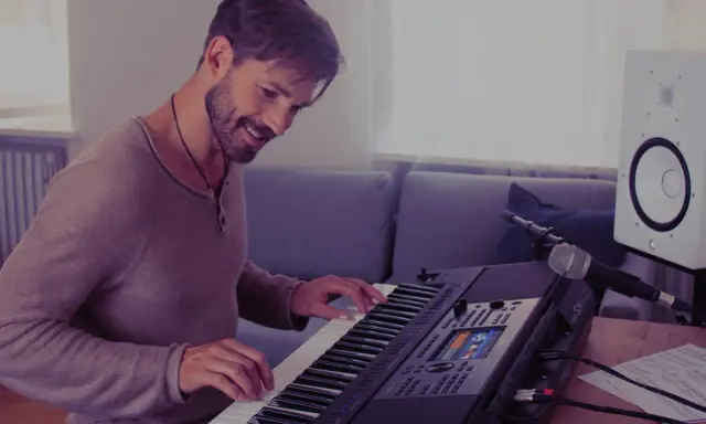 Uśmiechnięty mężczyzna gra na keyboardzie Yamaha, korzystając z aplikacji yamaha flowkey. Obok stoi mikrofon i głośnik.